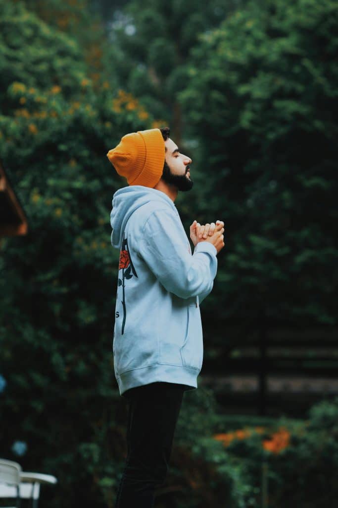A man in a beanie and hoodie deeply praying outside in a lush green forest.