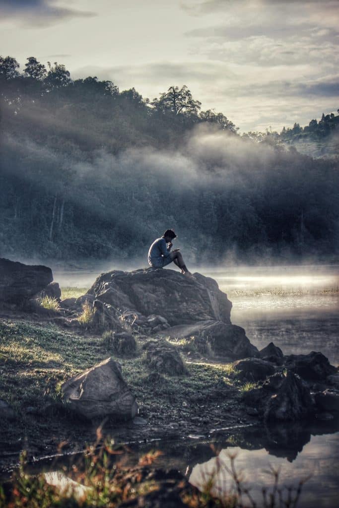 A lone figure reflects on a rock by a misty lake in Rancabali amidst tranquil surroundings.